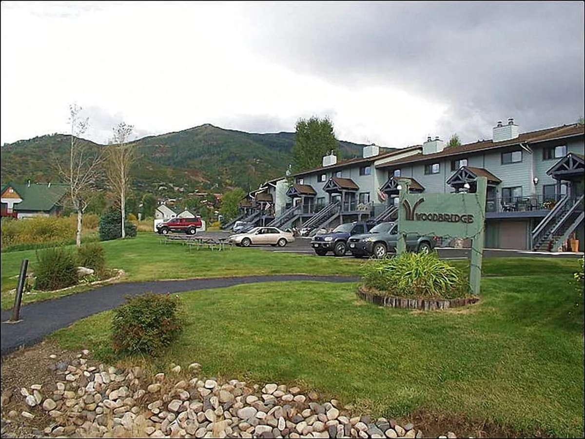 A view of the 'Woodbridge' community sign, with a well-maintained lawn, parked cars, and a mountainous backdrop. The image sets the context of a welcoming and peaceful neighborhood, hinting at community amenities and ease of access for guests.