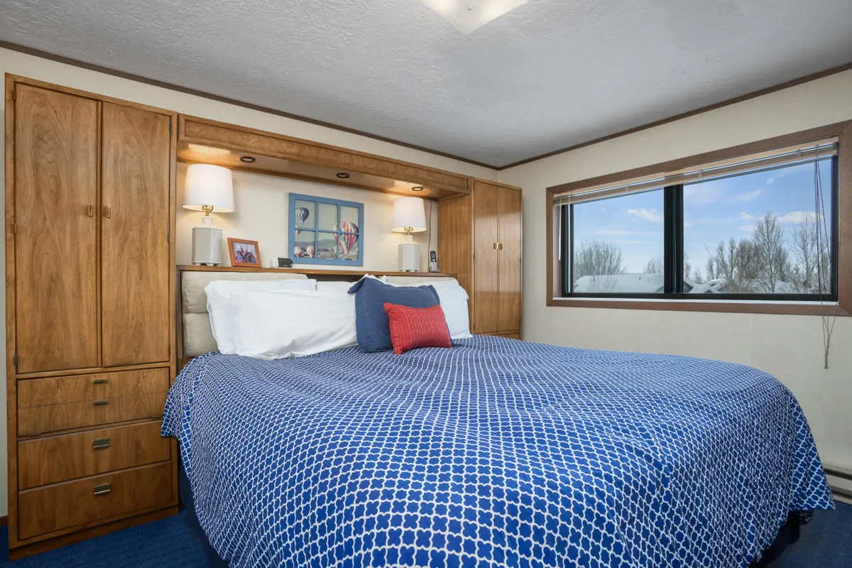This image showcases a cozy bedroom with a bed covered in blue patterned bedding, wooden cabinetry, framed pictures on the wall, and a window that brings in natural light and offers a calming view. The setup suggests a comfortable and restful space, perfect for relaxation and personal downtime.