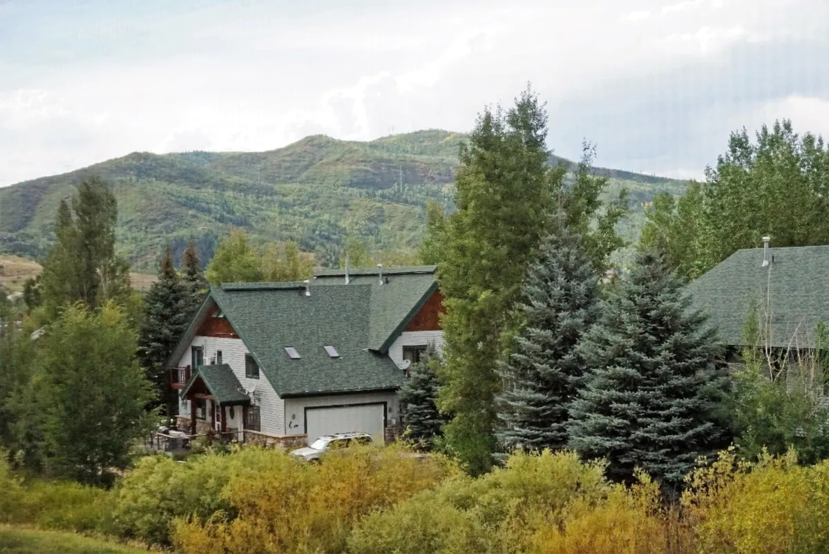 A wide exterior view of the property set against a mountain backdrop, framed by lush trees. The house stands as a gateway to the outdoors, suggesting close proximity to trails and nature. The landscape evokes the spirit of adventure and river recreation.