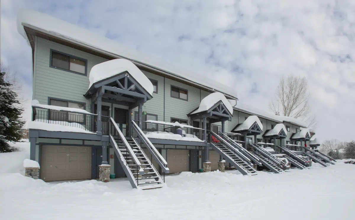 Snow-covered roofs and staircases lead to welcoming front doors, creating a cozy, inviting winter scene. The environment suggests a warm retreat after outdoor adventures, with the promise of fireside chats and family gatherings.