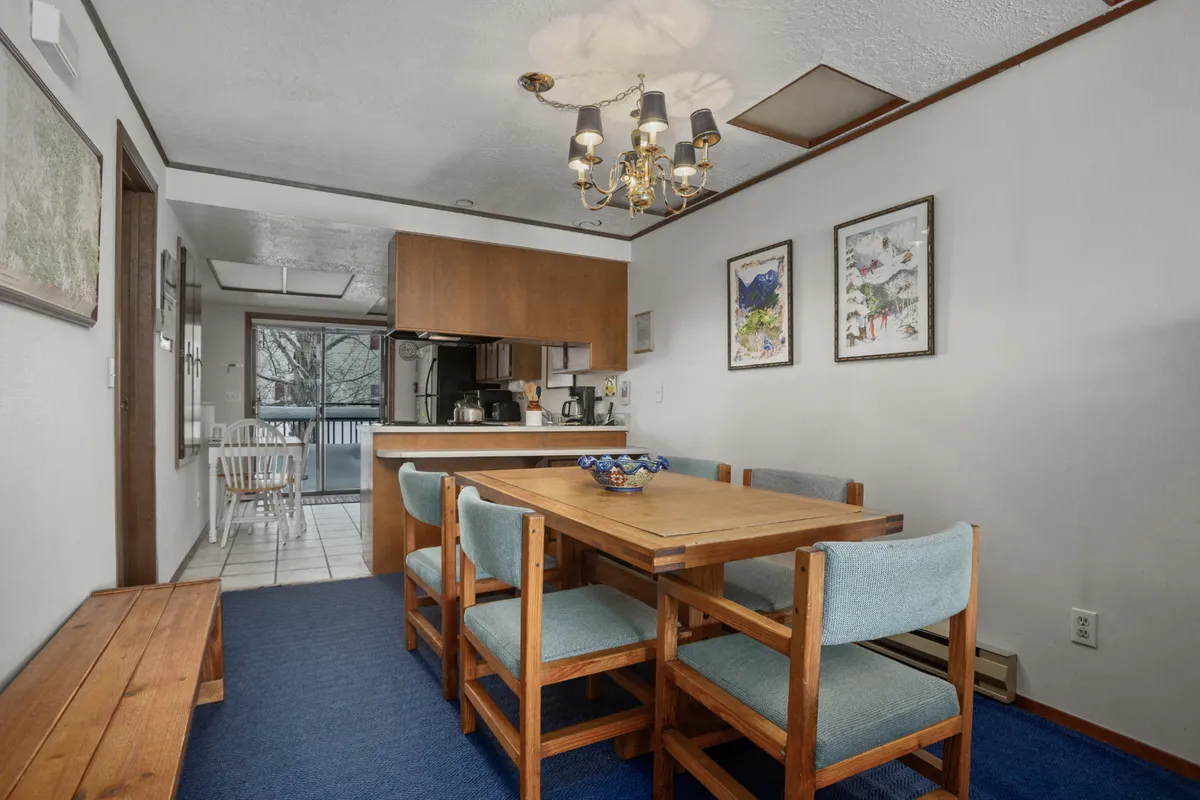 This image offers a view of a wooden dining table under a chandelier, with framed artwork on the wall and the kitchen area in the background. The setup encourages both cooking and togetherness, showcasing the kitchen's proximity to the dining space.