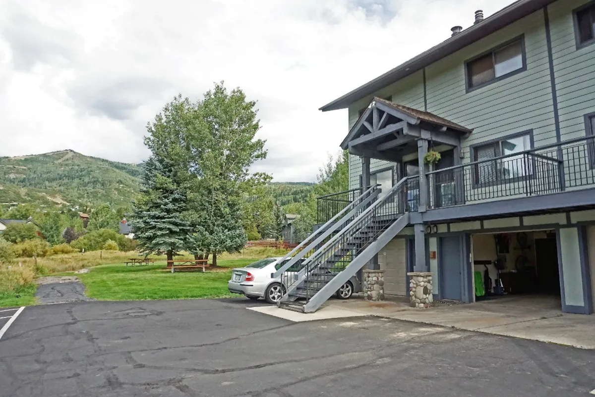 The image showcases the condo's exterior, featuring a welcoming staircase, a parked vehicle hinting at easy arrival, lush greenery surrounding the property, and a dramatic mountain backdrop. The scene is set at sunset, with warm light washing over the landscape, and plenty of open space for outdoor activities such as family picnics or stargazing.