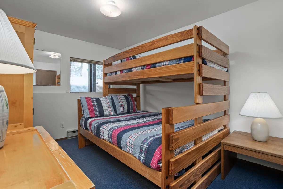 A charming bedroom with a bunk bed dressed in plaid bedding, a desk lamp for reading, and a window that adds brightness to the space. The setup is playful yet practical, ideal for kids or groups, and suggests flexibility in sleeping arrangements.