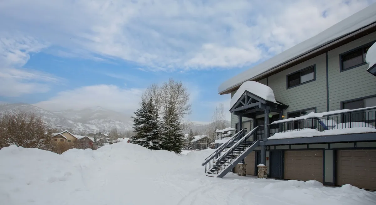 A snow-covered landscape frames modern architecture and tall trees, evoking the peacefulness of winter in Steamboat. The scene suggests activities like skiing, snowboarding, and cozy gatherings in a mountain setting.